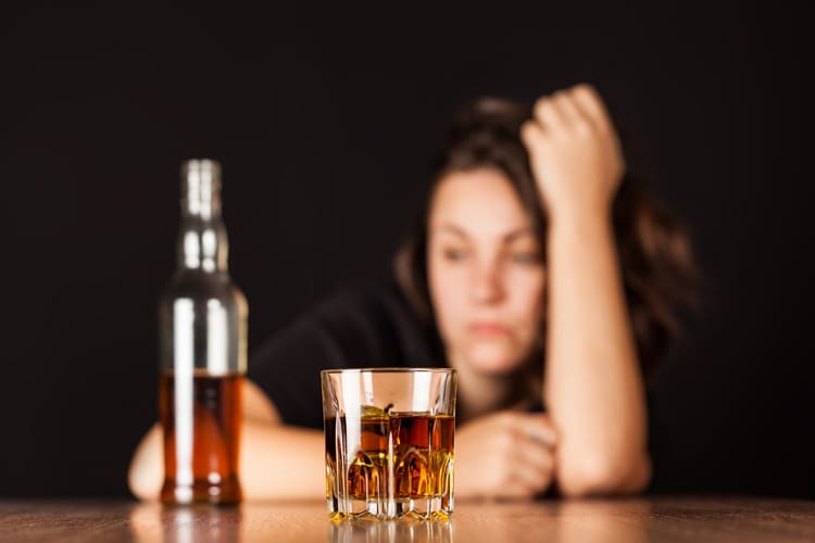 upset looking young woman behind a bottle and full glass of liquor - alcohol addiction
