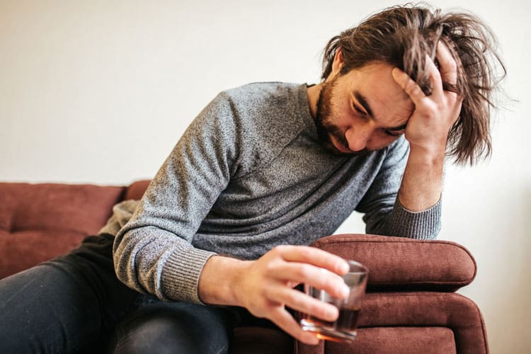 Signs of Addiction, middle age man with long hair and a beard looking defeated, holding a glass of liquor while on the couch at home - addiction signs