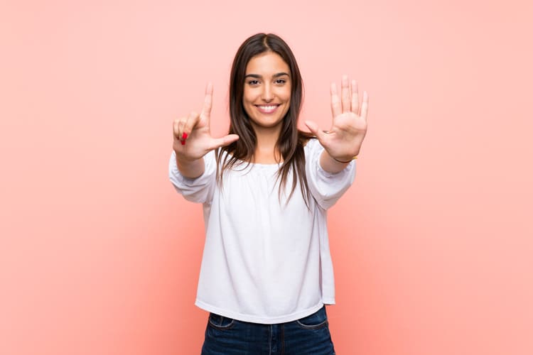 pretty young woman signing the number seven with her hands against a pink background - myths