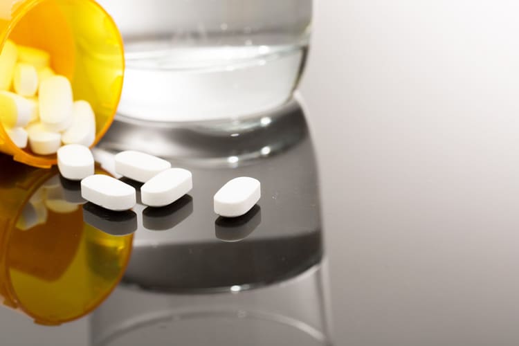 closely cropped shot of pain pills spilling out of prescription bottle next to a glass of water - opioid addiction