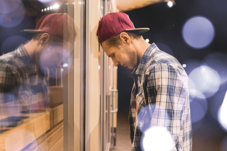 attractive young man in a baseball cap and flannel shirt leans his head against the outside window of a shop - dry drunk syndrome
