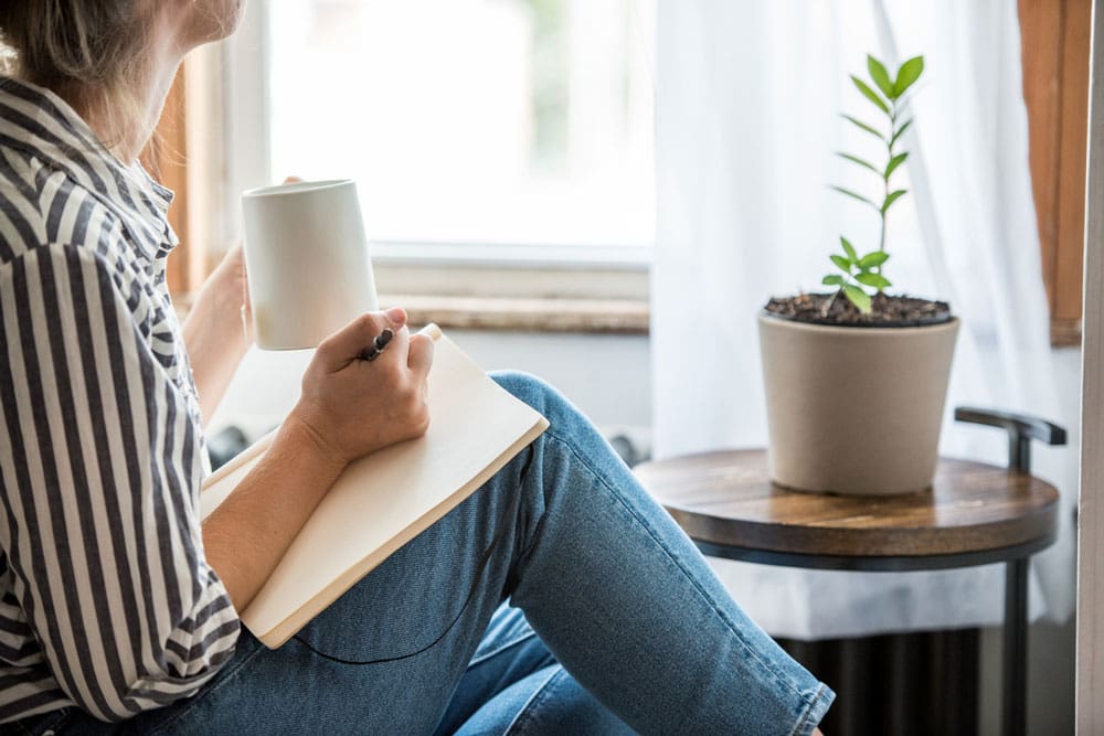 woman sitting with coffee and a journal by her window