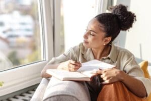 A young woman with curly hair in a bun sits comfortably by a large window, holding an open journal and pen. She looks thoughtfully outside while pausing in her writing. She wears an oversized beige shirt and orange pants, appearing calm and reflective in the cozy, sunlit space.