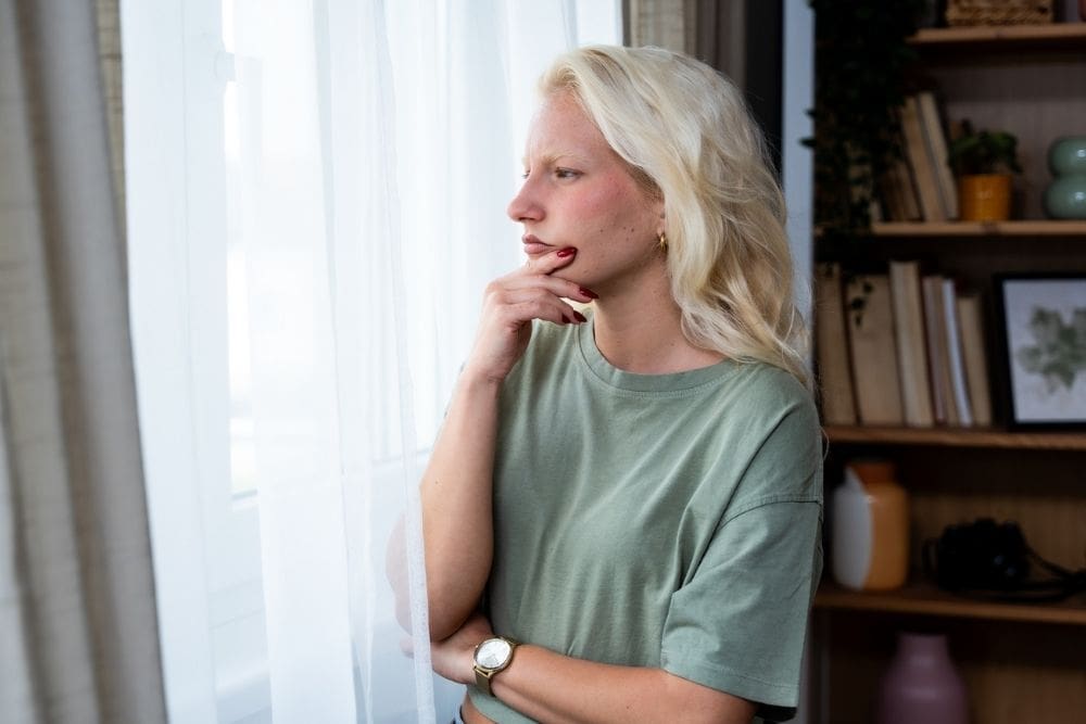 Young woman with long blonde hair wearing a green shirt, standing indoors by a window with sheer white curtains, looking outside thoughtfully with her hand resting on her chin.