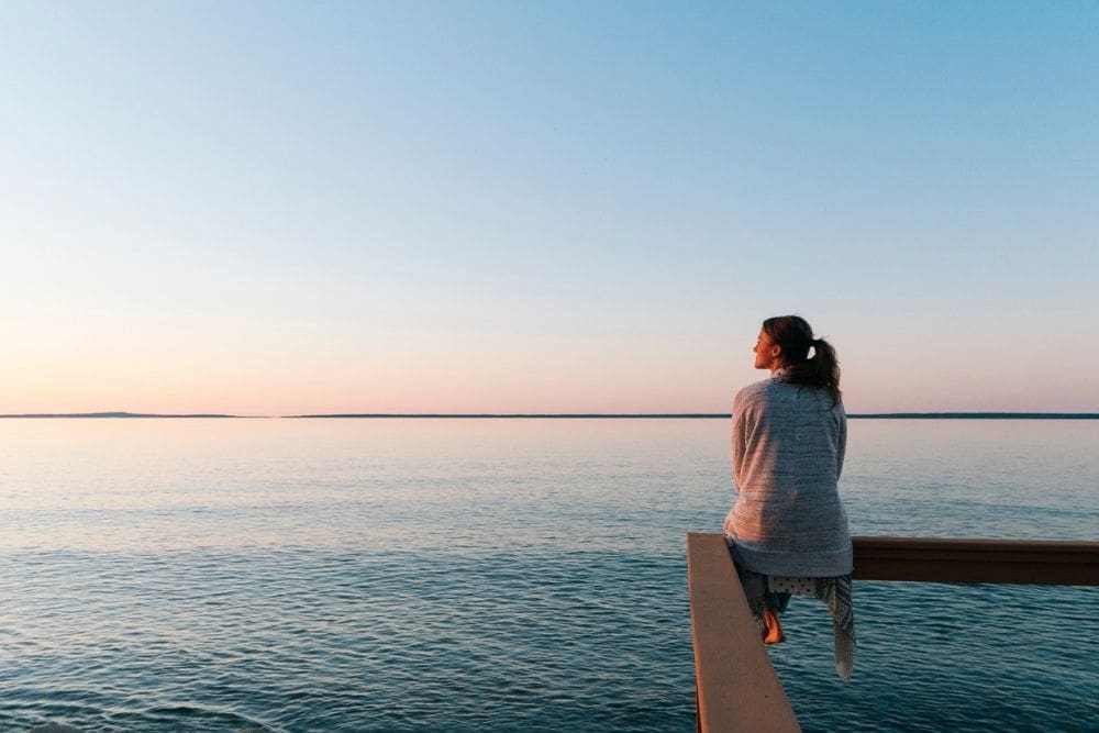 Woman sitting peacefully on a wooden railing overlooking a calm body of water at sunset, reflecting in a serene and contemplative moment.