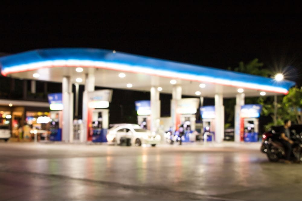 A blurry nighttime view of a brightly lit gas station with multiple fuel pumps and vehicles, creating a sense of activity and movement.