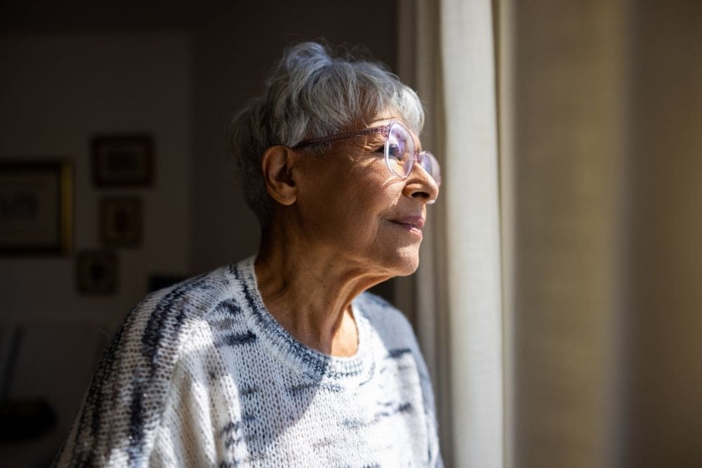 An older woman with short gray hair and glasses gazes thoughtfully out a window, sunlight illuminating her face as she stands in a cozy, softly lit room.