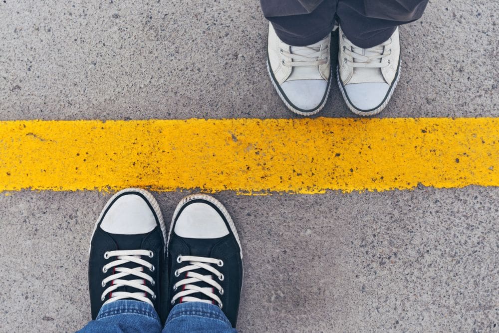 Two pairs of sneakers face each other on a concrete surface, separated by a bold yellow line, symbolizing personal boundaries.