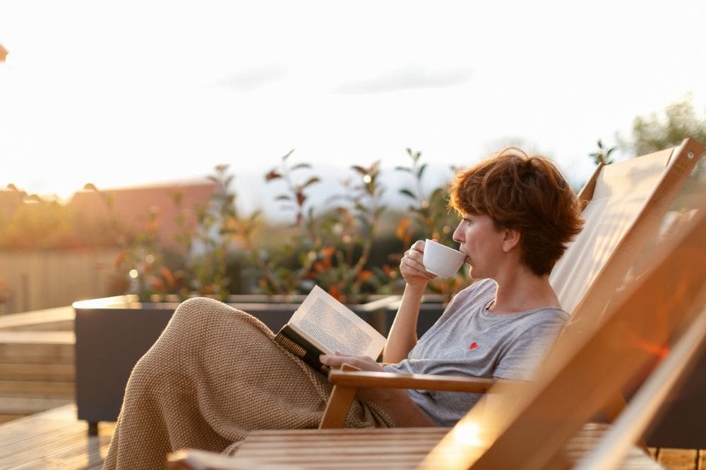 Woman relaxing on a lounge chair outdoors at sunset, wrapped in a blanket, sipping from a cup and reading a book, enjoying a peaceful moment.
