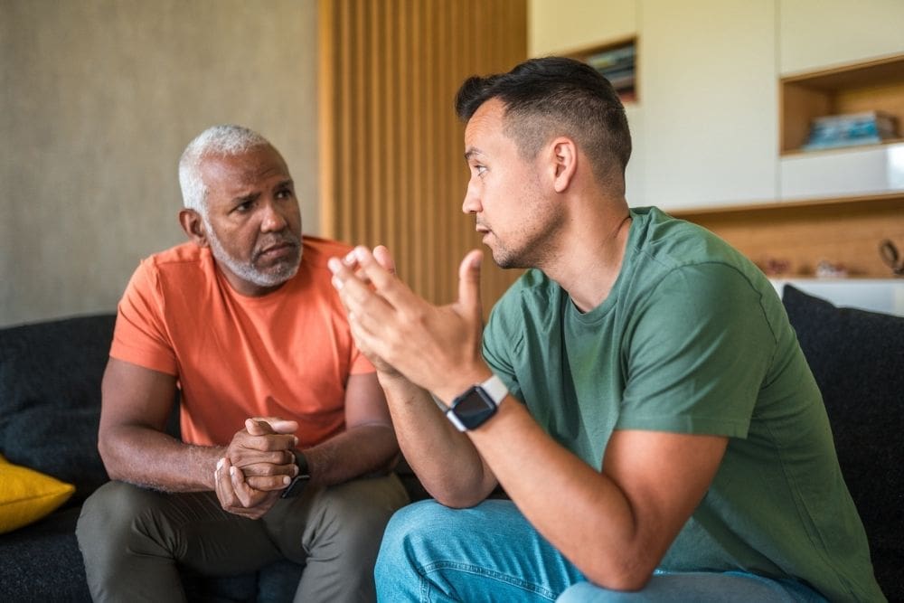 Two middle-aged men of diverse racial backgrounds engage in an intimate conversation within a living room