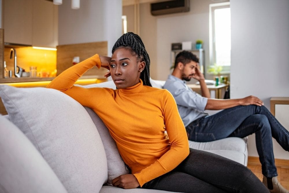 A woman sits on a couch looking upset while a man sits in the background with his head in his hand, symbolizing the strain of a toxic relationship.