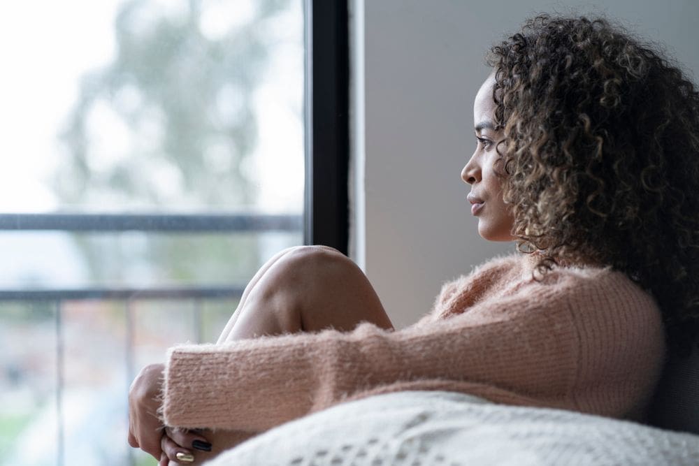 A woman sits indoors by a window with her knees pulled close, gazing outside thoughtfully, suggesting reflection, sadness, or emotional struggle