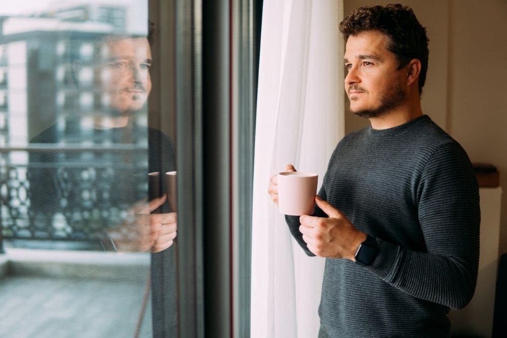Man holding a mug and looking out of a window with a thoughtful expression.