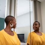Positive Self talk (1000 x 667 px) Smiling woman in a yellow shirt looking at her reflection in the mirror, symbolizing confidence and positive self-talk in recovery.
