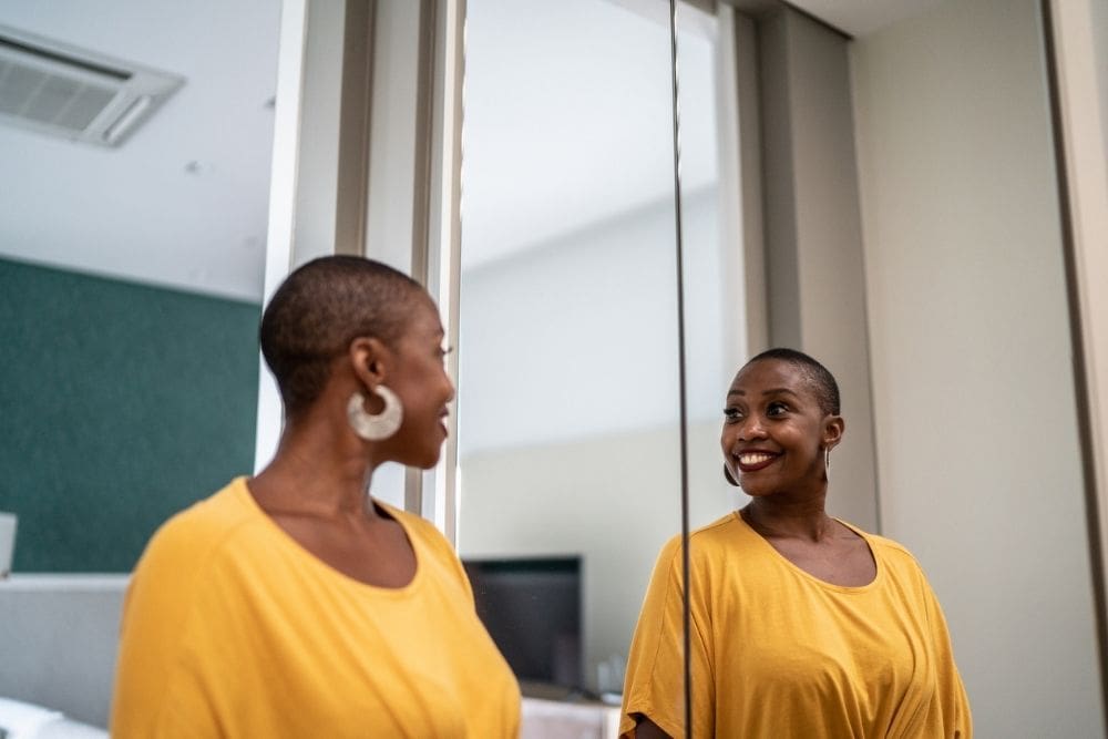 Smiling woman in a yellow shirt looking at her reflection in the mirror, symbolizing confidence and positive self-talk in recovery.