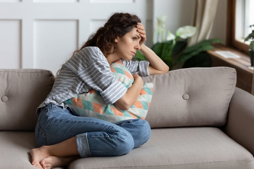 Woman sitting on a couch hugging a pillow, looking worried and deep in thought, suggesting anxiety or emotional stress.