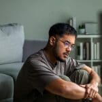 Dual Diagnosis(1000 x 667 px) A man sits on the floor indoors with his arms wrapped around his knees, looking thoughtful and concerned.