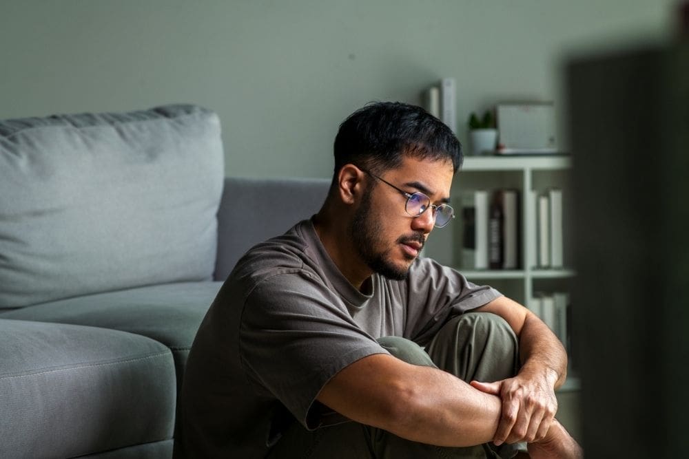 A man sits on the floor indoors with his arms wrapped around his knees, looking thoughtful and concerned.