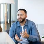A man sits in a bright, modern office space, speaking with a therapist. He gestures with his hands while engaging in conversation, showing an open and attentive expression.