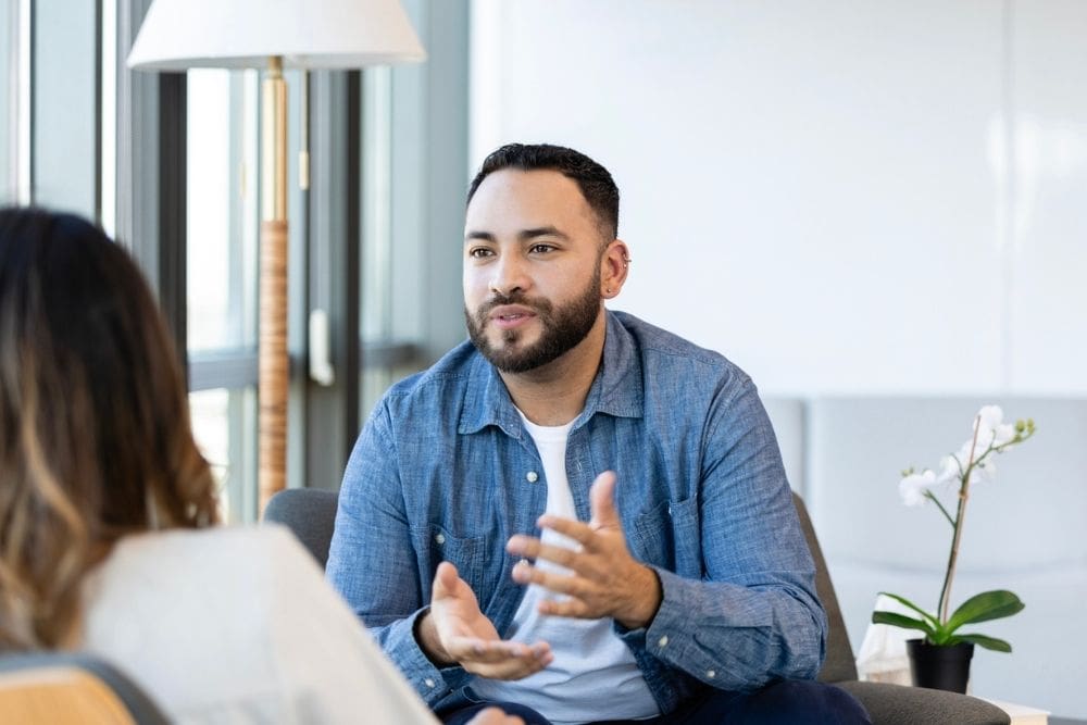A man sits in a bright, modern office space, speaking with a therapist. He gestures with his hands while engaging in conversation, showing an open and attentive expression.