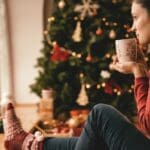Holiday Rest (1000 x 667 px) A woman sitting by a window with a warm drink, wearing cozy holiday socks and looking out thoughtfully, with a decorated Christmas tree in the background.