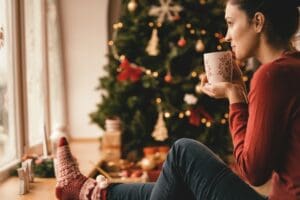 A woman sitting by a window with a warm drink, wearing cozy holiday socks and looking out thoughtfully, with a decorated Christmas tree in the background.