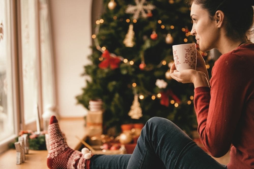 Holiday Rest (1000 x 667 px) A woman sitting by a window with a warm drink, wearing cozy holiday socks and looking out thoughtfully, with a decorated Christmas tree in the background.