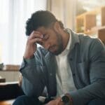 A man sitting indoors with his head resting in his hand, appearing overwhelmed or distressed in a quiet, softly lit room.