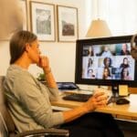 A woman sits at a desk holding a mug while attending a virtual group therapy session on a computer in a home office setting.