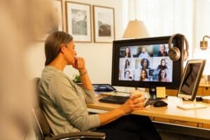 A woman sits at a desk holding a mug while attending a virtual group therapy session on a computer in a home office setting.