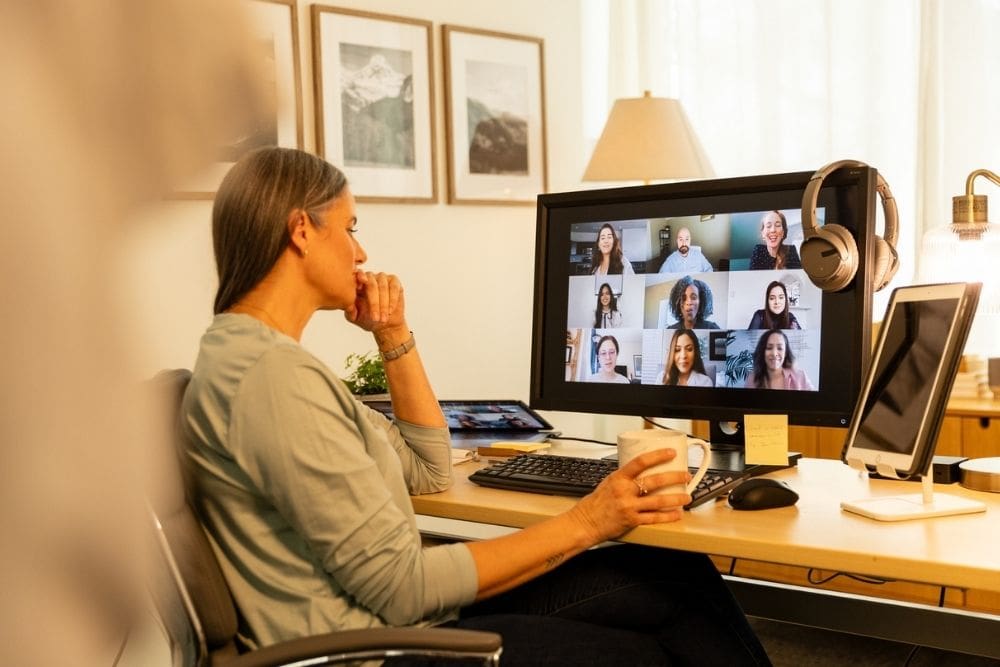 Virtual IOP (1000 x 667 px) A woman sits at a desk holding a mug while attending a virtual group therapy session on a computer in a home office setting.