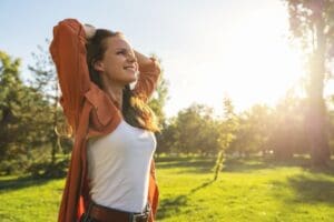 How Addiction detox Can Save Your Life - A woman smiles joyfully with hands behind her head, enjoying sunlight in a green park. She's wearing an orange shirt and a white tank top. The scene feels serene and uplifting.