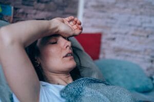 Anabranch - What Happens in Alcohol Detox - Edited. A woman lays in bed with her hand ove rher forehead checking her temperature. She has her eyes closed and is presenting as being sick.